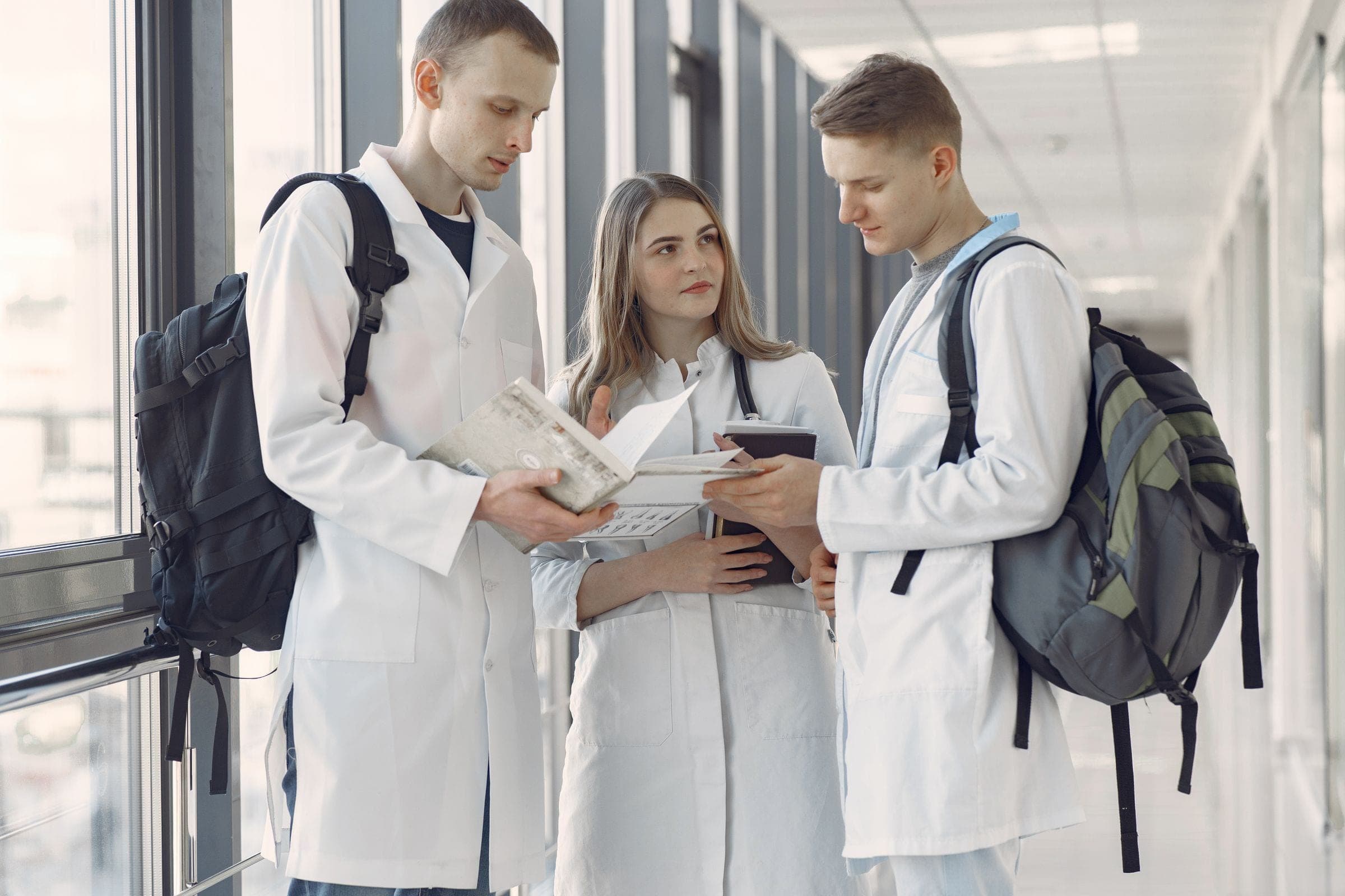 Three early-career clinicians in white coats comparing notes in a bright hospital corridor.