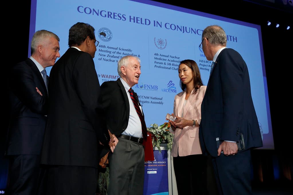 Federation leaders in conversation on stage at the World Congress, before a screen naming the partner organisations, including the IAEA.